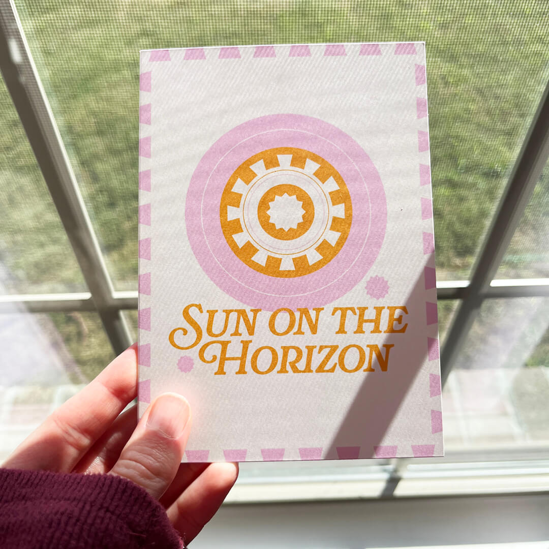 Hand holding a pink and orange greeting cards that reads "Sun on the horizon" with an abstract sun motif